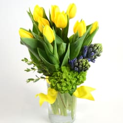 Yellow tulips arranged in a clear glass vase with green and purple flowers
