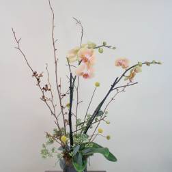 Tall orchid arrangement in a ribbed metal vase with branches and round yellow accents