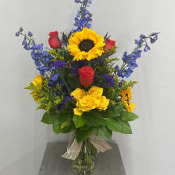 Tall bouquet of sunflowers, red and yellow roses, and blue flowers in a glass vase