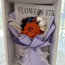 Boxed arrangement with orange rose, blue blooms, and cotton in lavender wrap with white ribbon