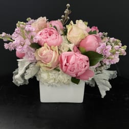 Low arrangement of pink peonies and blush roses in a white square vase with soft white flowers.