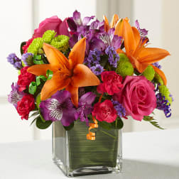 Mixed bouquet of orange lilies, pink roses, and purple flowers in a glass vase