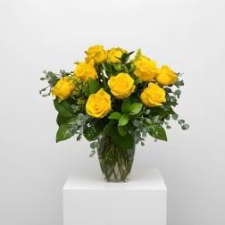 Arrangement of bright yellow roses in a clear glass vase with lush filler accents.