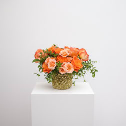 Low arrangement of orange and peach roses in a gold textured glass vase with trailing accents