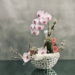 Pink orchids arranged in a white decorative bowl with small gift items