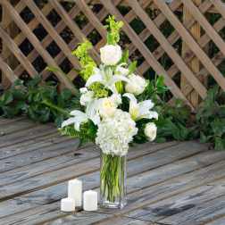 White floral arrangement in a tall glass vase with three candles nearby