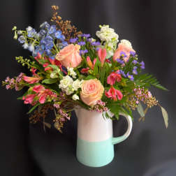 Mixed bouquet of pastel roses, blue delphinium, and purple daisies in a two-tone pitcher