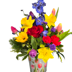 Colorful arrangement of yellow daffodils, red roses, tulips and blue delphinium in a printed metal bucket.