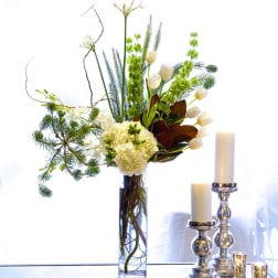 Tall white floral arrangement in a glass vase beside silver candle holders