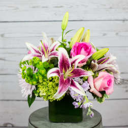 Compact arrangement of pink lilies and roses with green accents in a square glass vase.