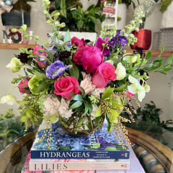 Low glass vase of bright pink roses and mixed purple and green flowers on a stack of flower books.
