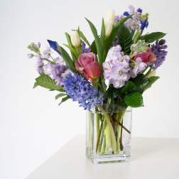 Mixed bouquet of pink roses, purple stock, blue blooms, and white tulips in a clear glass vase