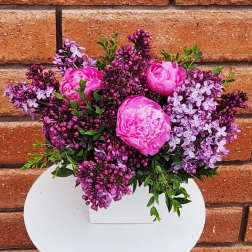 Low arrangement of bright pink peonies and purple lilacs in a white square vase.