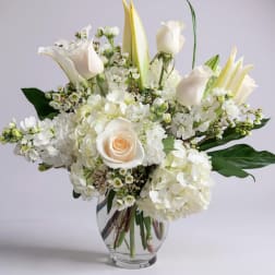 White floral arrangement in a clear glass vase with roses, calla lilies, and hydrangeas