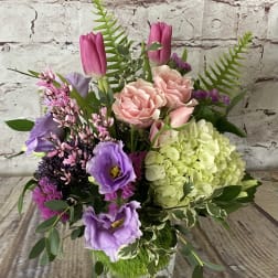 Mixed arrangement of pink roses, purple tulips, lavender lisianthus, and green hydrangea in a glass cube vase