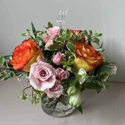 Short glass vase arrangement with orange-yellow and pink roses and variegated foliage
