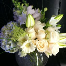 Low round arrangement of white roses, lilies, hydrangeas, gerbera daisies, and snapdragons in a clear glass bowl vase