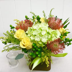 Low arrangement of green hydrangea, yellow roses, and red pincushion protea in a square vase with a small votive candle.