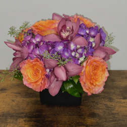 Low arrangement of orange roses, purple hydrangeas, and mauve orchids in a black square vase