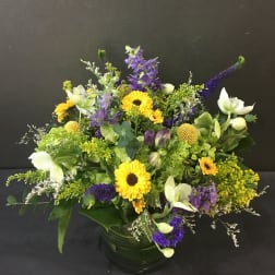 Low mixed arrangement of yellow and purple flowers in a round clear glass vase