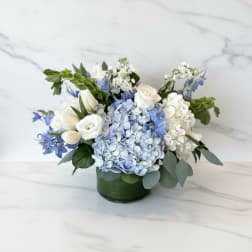 Low arrangement of white roses, tulips, and blue hydrangeas in a green glass cylinder vase