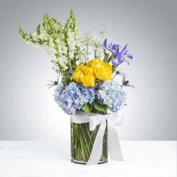 Tall arrangement with yellow roses, blue hydrangeas, white snapdragons and purple iris in a glass vase with white ribbon