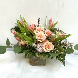 Peach roses and pink tulips arranged in a low clear glass cube vase with white filler flowers.