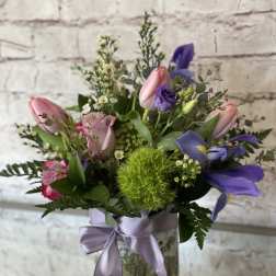 Mixed bouquet of pink tulips, roses, and purple iris in a glass vase with lavender ribbon