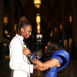 Couple in formal attire exchanging a boutonniere in a dim hallway