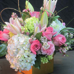 Compact arrangement of pink roses, white hydrangeas, and lilies in a gold cube vase