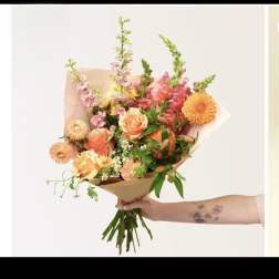 Handheld bouquet of peach roses, dahlias, and mixed flowers wrapped in brown paper