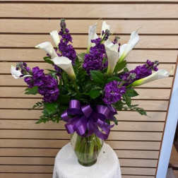 Purple flowers and white calla lilies in a glass vase with a purple ribbon