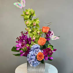 Tall arrangement of green and purple orchids, orange roses, and blue hydrangea in a square vase with butterfly picks
