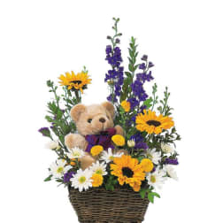 Teddy bear in a wicker basket surrounded by yellow and white daisies and purple flowers.