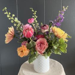 Mixed bouquet of pink, peach, yellow, and purple flowers in a white vase
