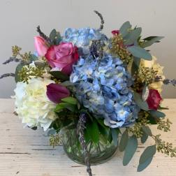 Low glass vase with blue hydrangeas, white blooms, and pink roses in a rounded arrangement.