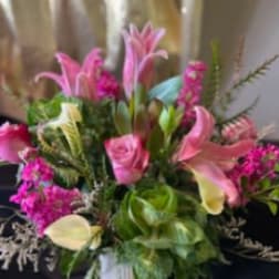 Pink lilies, roses, and pale calla lilies arranged with bright filler flowers in a white vase