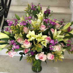 Large mixed bouquet of pale yellow lilies, pink roses, and purple flowers in a clear glass vase