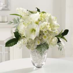 White amaryllis, roses, and small daisies in a clear glass vase with glass stones and ribbon.