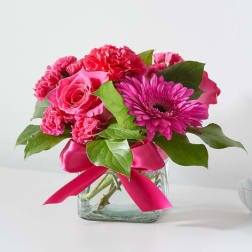 Bright pink roses, carnations, and a gerbera daisy in a square glass vase with a pink ribbon bow.