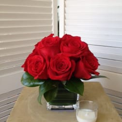 Red roses arranged in a square glass vase with a small candle nearby
