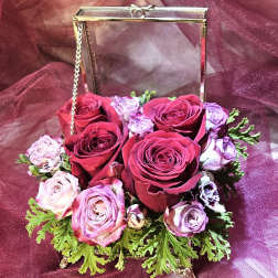 Compact arrangement of red and lavender roses in a gold metal and glass box on a burgundy fabric background