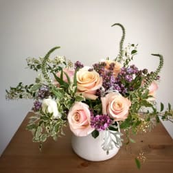 Low arrangement of peach roses and purple blossoms in a white ceramic vase on a wooden table
