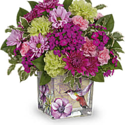 Compact arrangement of purple mums and green and pink carnations in a square hummingbird vase