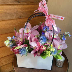 Pink orchid arrangement in a white gift bag with a heart ribbon