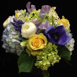 Compact bouquet of yellow roses, white and purple flowers in a glass vase against a dark background