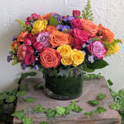 Bouquet of orange, yellow, pink, and lavender roses with purple asters in a clear cylinder vase.