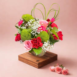 Pink and red floral arrangement in a glass vase on a wooden base