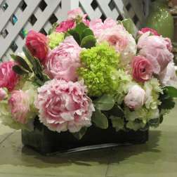 Low arrangement of pink peonies, ranunculus, and white and green hydrangeas in a rectangular glass vase
