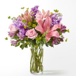 Pink lilies, roses, and purple flowers arranged in a clear glass cylinder vase